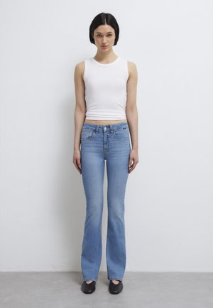 Frau mit schwarzen Haaren steht vor einer weißen Wand, trägt ein ärmelloses weißes Top, hellblaue Jeans und schwarze Schuhe und schaut nach vorne.