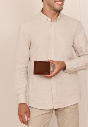 Man in beige shirt and pants holds a brown leather wallet at chest level, smiling and looking to his right.
