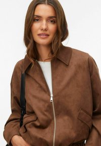 Smeđa jakna od brušene kože s ovratnikom, prednjim patentnim zatvaračem i bočnim džepovima. Ima ležeran kroj i teksturiranu površinu.