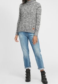 Pull à col roulé tricoté gris et noir, associé à un jean bleu clair retroussé et des bottines noires cloutées. Étiquette visible sur le pull.