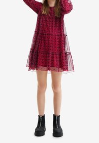 Vestido rojo con volantes y un patrón en espiral, mangas largas transparentes y un escote redondeado. Llevado con botas negras de tobillo con cremalleras.