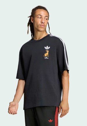 Mann mit Dreadlocks, der ein schwarzes Adidas-Shirt mit Cartoon-Hund-Grafik und weißen Streifen an den Ärmeln trägt, kombiniert mit schwarzen Hosen mit roten Streifen.