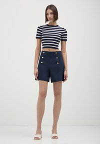 Donna che indossa pantaloni corti navy a vita alta con bottoni dorati, un top a righe navy e bianche crop, e sandali bianchi con tacco, in piedi su un pavimento bianco.