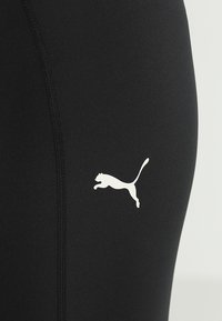 Leggings neri realizzati in un tessuto liscio e elasticizzato, con un logo Puma bianco sulla gamba sinistra e dettagli delle cuciture visibili.