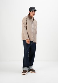 Man wearing beige jacket with brown collar, dark blue cuffed jeans, brown sneakers, and black cap, standing against white background.