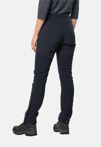 Pantalons de randonnée extensibles bleu marine au design ajusté, avec des évents à zip au niveau des chevilles et une texture lisse. Associés à des bottes de randonnée grises.