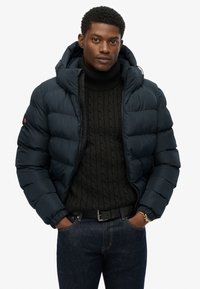 Marineblauwe pufferjack met capuchon, voorzien van een gewatteerd ontwerp, elastische manchetten en een rits aan de voorkant. Draagt over een zwart kabelgebreid sweater.