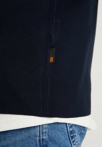 Marinblå tröja med ribbat mönster och en sidoseamsmärke med en svart rektangel och en orange kvadrat. Vit skjorta under, i kombination med denim.