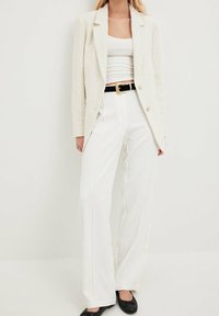 Valkoinen teksturoitu bleiseri, jossa on kaksi kultaa nappia, yhdistetty valkoisiin housuihin ja mustaan vyöhön. Mustat litteät kengät täydentävät asua.