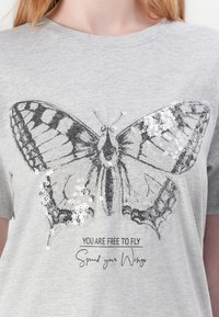 T-shirt gris à manches courtes avec un graphique de papillon en contour noir et des sequins argentés, accompagné des textes "VOUS ÊTES LIBRE DE VOLER" et "Ouvrez vos ailes."