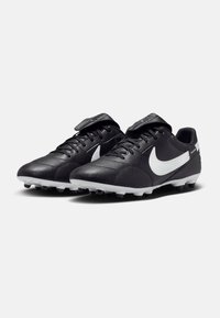 Crampons de football en cuir noir avec des accents blancs, bout arrondi et surface texturée, dotés de lacets et d'une sangle velcro pour un meilleur maintien.