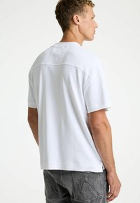 Camiseta blanca de manga corta con un corte relajado, que presenta un cuello redondo y detalles sutiles de costuras texturizadas. Combinada con pantalones vaqueros grises.