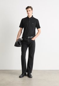 Black short-sleeved polo shirt with three buttons and a logo on the chest, paired with black jeans and black shoes, holding a black bag.