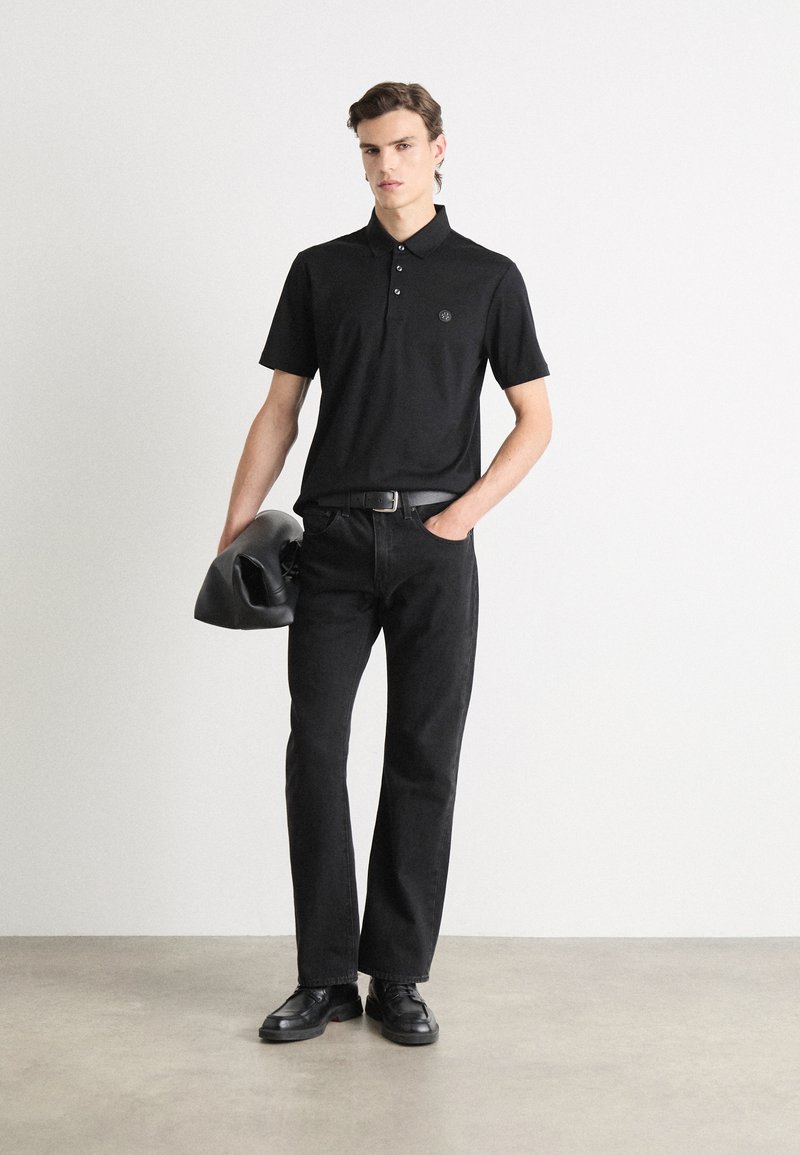 Schwarzes Polohemd mit kurzen Ärmeln, drei Knöpfen und einem Logo auf der Brust, kombiniert mit schwarzen Jeans und schwarzen Schuhen, hält eine schwarze Tasche.