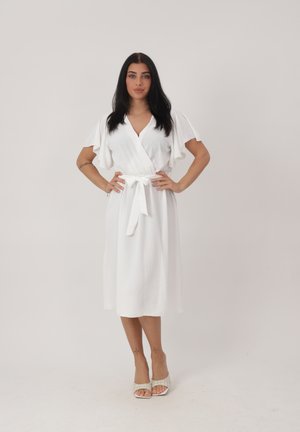 Kvinde med langt mørkt hår iført en hvid slå-om-kjole med flagrende ærmer og bundet talje, stående med hænderne på hoften mod en ensfarvet baggrund.