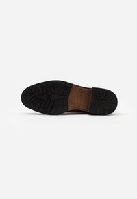 Semelle en caoutchouc noir avec motif de bande de roulement prononcé, comprenant une semelle intermédiaire marron et un marquage en relief. Le design est plat et reste flexible.