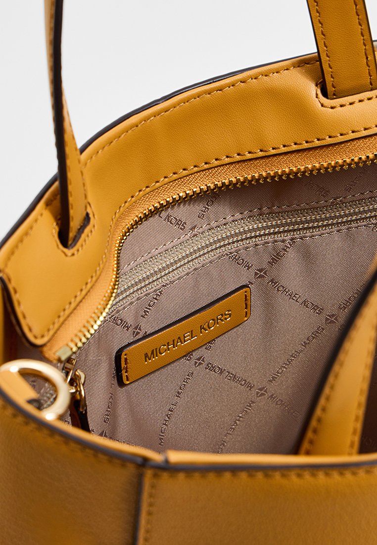 Intérieur d'un sac fourre-tout en cuir jaune avec une poche zippée et une doublure en tissu arborant le logo de la marque. Des accents en métal doré sont visibles.