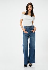 Weißes, schulterfreies Rippenoberteil kombiniert mit hoch taillierten, weit geschnittenen blauen Jeans, die mit einem Knopf- und Reißverschlussverschluss ausgestattet sind, sowie schwarzen, spitzen High Heels.