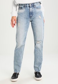 Jeans de mezclilla de tiro alto en azul claro, con un corte relajado y detalles desgastados en los muslos, combinados con zapatos negros tipo slip-on.