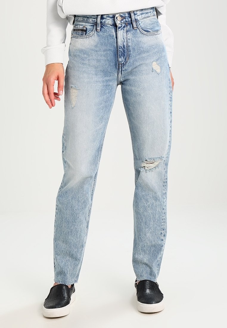 Jeans de mezclilla de tiro alto en azul claro, con un corte relajado y detalles desgastados en los muslos, combinados con zapatos negros tipo slip-on.