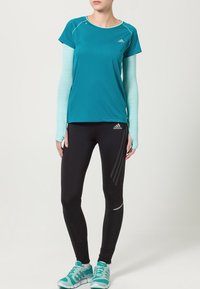 Teal lyhythihainen paita, jossa on vaaleansiniset pitkät hihat, mustat legginsit harmailla yksityiskohdilla ja teal-väriset urheilukengät; sisältää Adidas-logon.
