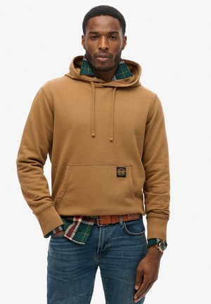 Felpa marrone realizzata in un tessuto morbido, con tasca frontale e lacci, indossata sopra una camicia a quadri verde e rossa, abbinata a jeans blu.