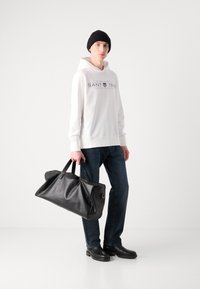 Borsa da viaggio in pelle nera con due manici, caratterizzata da una texture liscia e design pieghevole, tenuta da una persona in felpa bianca e jeans blu.