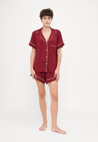 Βουργουνδί σετ πιζάμας που περιλαμβάνει κοντομάνικο πουκάμισο με κουμπιά και λευκή διακόσμηση, καθώς και ασορτί σορτσάκι. Λείο ύφασμα, άνετη εφαρμογή.