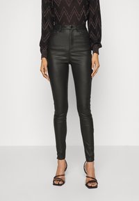 Svarta höga fusk läderbyxor med smal passform, med en slät textur och minimal detaljering, i kombination med svarta sandaler med remmar.