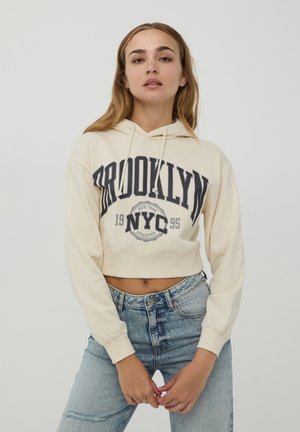 Felpa beige tagliata con grafica "BROOKLYN NYC 1995" in blu scuro. Presenta un cappuccio con cordino e polsini a costine. Abbinata a jeans a vita alta.