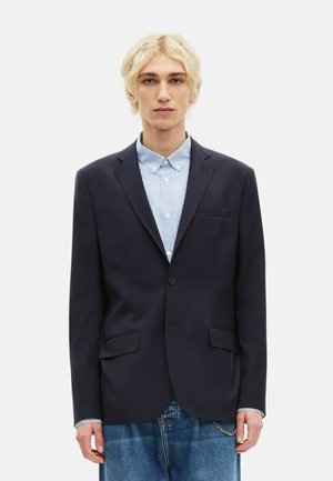 Młoda osoba o krótkich blond włosach, ubrana w ciemnogranatową marynarkę na jasnoniebieskiej, pasiastym koszuli z guzikami oraz luźnych niebieskich dżinsach.