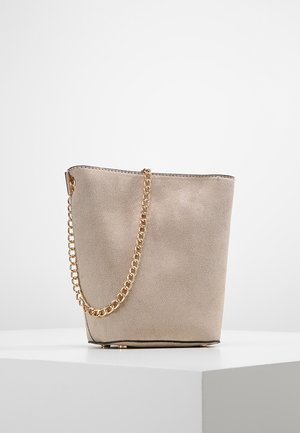 Beige suède buckettas met een gouden kettingriem, zwarte voering en een stevige, platte bodem, staand op een wit oppervlak.