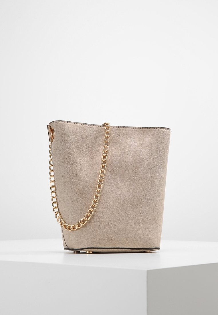 Sac seau en daim beige avec une bandoulière chaîne dorée, doublure noire et une base plate structurée, posé sur une surface blanche.