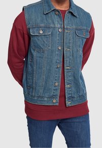 Denim ärmlös jacka med två bröstfickor, silverknappar, slitna detaljer, bärs över en vinröd långärmad sweatshirt.