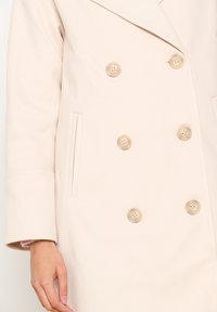 Dwurzędowy płaszcz w jasnym beżu wykonany z teksturowanej tkaniny; posiada duże beżowe guziki oraz boczne kieszenie, charakteryzując się luźnym krojem.