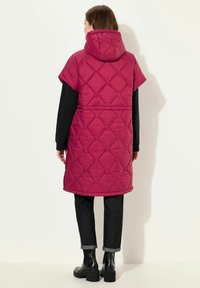 Dlouhá bezrukávová péřová bunda v živé magentové barvě s diamantovým prošíváním, nastavitelnou kapucí a bočními štěrbina, spárovaná s černými džíny a kotníkovými botami.