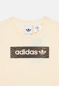 Jasno-kremowa koszulka z grafiką brązowych i czarnych pasków tygrysa oraz białym logo "adidas", z małą metką marki na kołnierzyku.