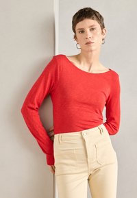 Vörös, hosszú ujjú felső széles nyakkivágással, könnyű anyagból készült, magas derekú bézs nadrággal párosítva, amely elülső zsebekkel rendelkezik.