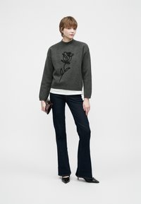 Jonge persoon die staat, gekleed in een donkergrijze trui met een zwart rozenontwerp, donkerblauwe flared jeans, zwarte puntige hakken, en een kleine handtas vasthoudend.