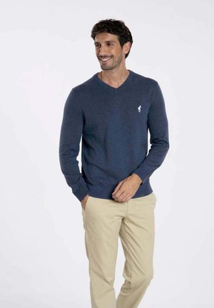 Pull en V bleu marine en tissu tricoté, avec un petit logo blanc sur la poitrine, associé à un pantalon beige.