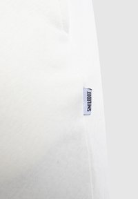 Weißer Baumwollstoff mit sichtbarer Textur, versehen mit einem kleinen blauen Etikett, auf dem der Text "SMILODOX" an der Seite gestickt ist.