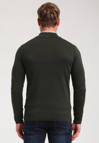 Pull vert foncé en tricot avec un col et des poignets côtelés, présentant une texture lisse. Coutures visibles le long des coutures et détail au dos.