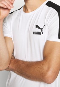 Vit atletisk t-shirt med svarta axeldetaljer, med en svart Puma-logotyp på bröstet. Slät tyg och korta ärmar.