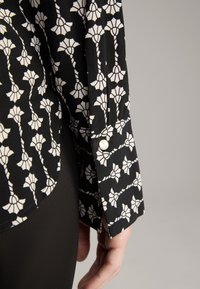 Black long-sleeve shirt with white floral pattern, featuring a buttoned cuff and smooth texture, close-up view of sleeve and hand.