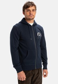 Námořnická mikina na zip vyrobená z měkké látky, s klokaní kapsou, kapucí se šňůrkou a tištěným logem na levé straně hrudi.