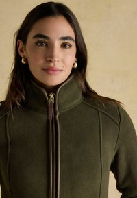 Woman with long dark hair wearing gold hoop earrings and an olive green fleece jacket with a front zipper against a beige background.