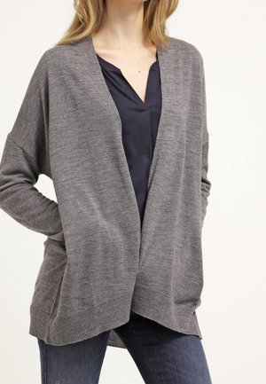 Femme portant un cardigan gris ample ouvert sur le devant, par-dessus un chemisier bleu foncé à col en V et un jean foncé, debout avec une main sur la hanche.