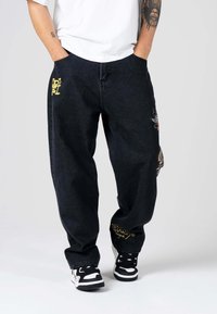 Pantalones de mezclilla negros con corte suelto, detalles de bordado amarillo en los bolsillos y un parche colorido en una pierna. Combinados con zapatillas negras y blancas.