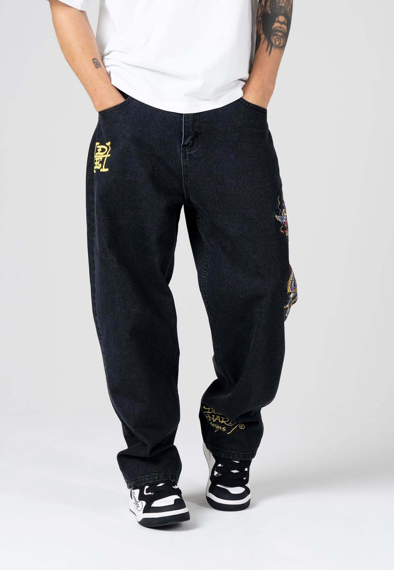 Pantalones de mezclilla negros con corte suelto, detalles de bordado amarillo en los bolsillos y un parche colorido en una pierna. Combinados con zapatillas negras y blancas.
