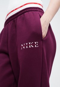 Bordo trenirke s bijelim Nike logom, koje imaju elastičnu traku u struku i bočne džepove. Tkanina je meka i blago teksturirana.